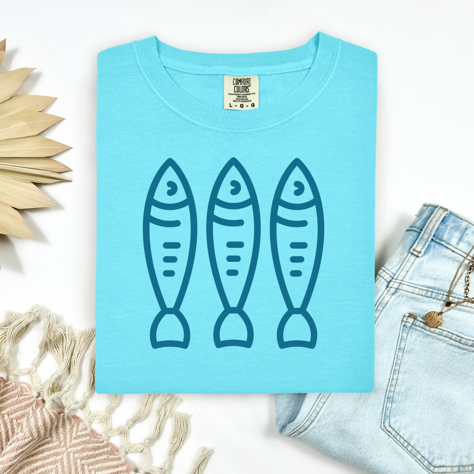 Comfort Colors 1717 Fisherman's Aesthetic t shirt with three sardines in a  one color graphic. Picture on a flay lay mockup with pair of blue jeans and blanket and a wicker sun shaped accessory.