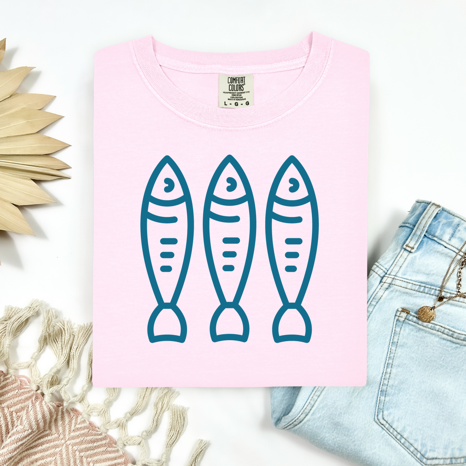 Comfort Colors 1717 Fisherman's Aesthetic t shirt with three sardines in a  one color graphic. Picture on a flay lay mockup with pair of blue jeans and blanket and a wicker sun shaped accessory.
