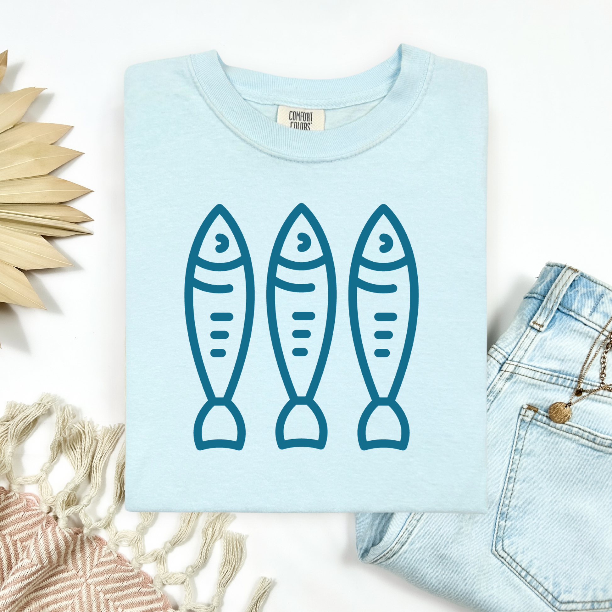 Comfort Colors 1717 Fisherman's Aesthetic t shirt with three sardines in a  one color graphic. Picture on a flay lay mockup with pair of blue jeans and blanket and a wicker sun shaped accessory.
