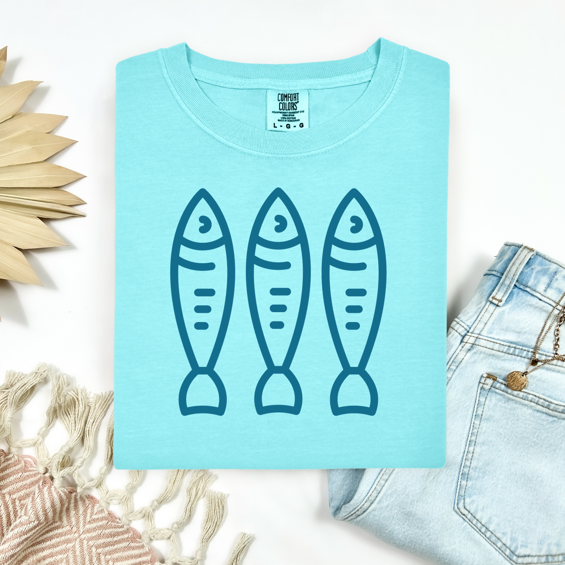 Comfort Colors 1717 Fisherman's Aesthetic t shirt with three sardines in a  one color graphic. Picture on a flay lay mockup with pair of blue jeans and blanket and a wicker sun shaped accessory.