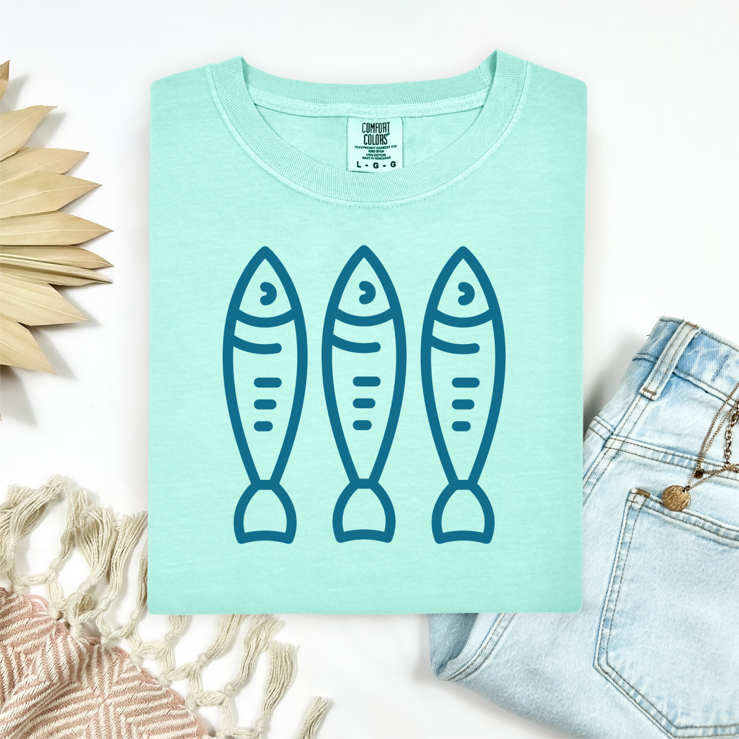 Comfort Colors 1717 Fisherman's Aesthetic t shirt with three sardines in a  one color graphic. Picture on a flay lay mockup with pair of blue jeans and blanket and a wicker sun shaped accessory.