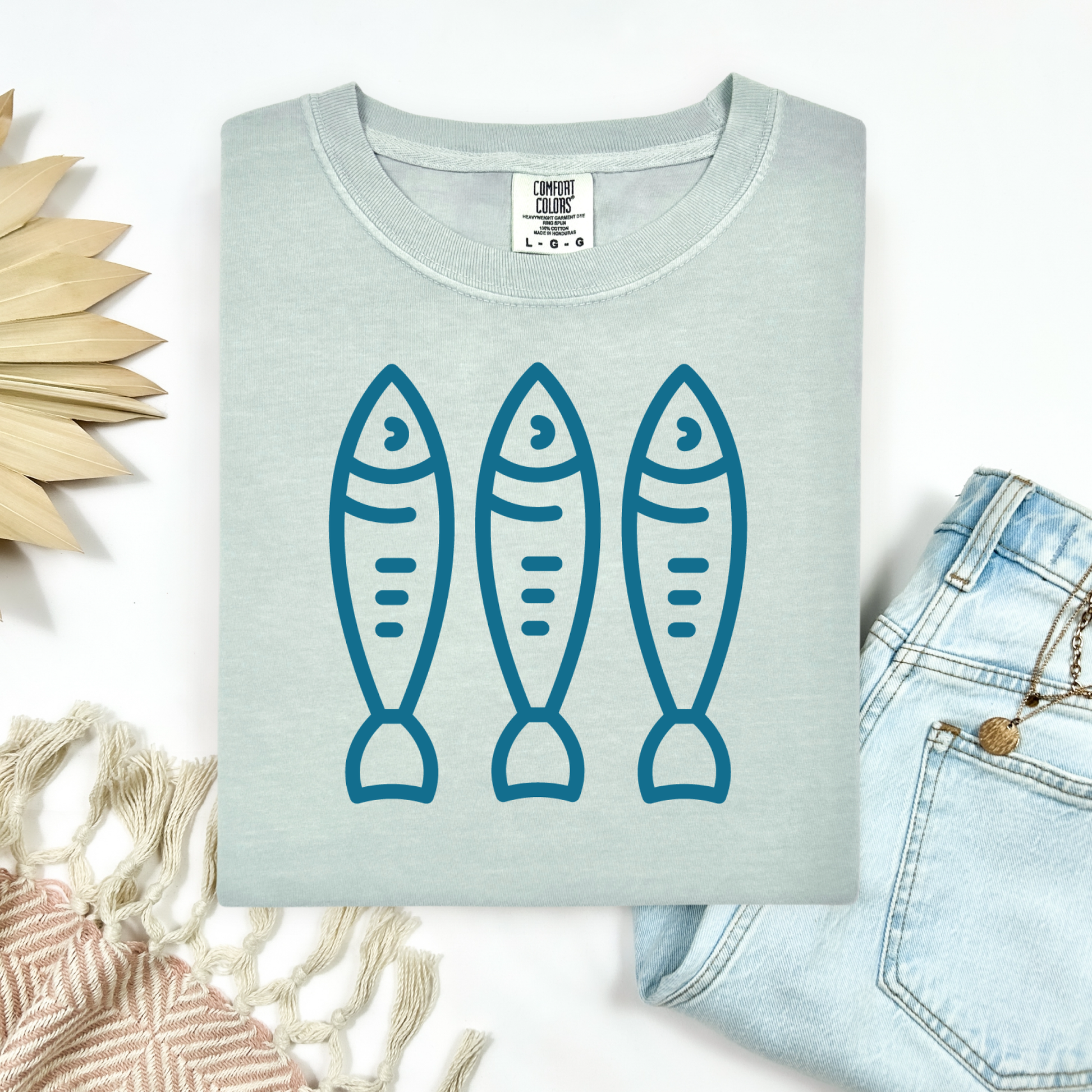 Comfort Colors 1717 Fisherman's Aesthetic t shirt with three sardines in a  one color graphic. Picture on a flay lay mockup with pair of blue jeans and blanket and a wicker sun shaped accessory.