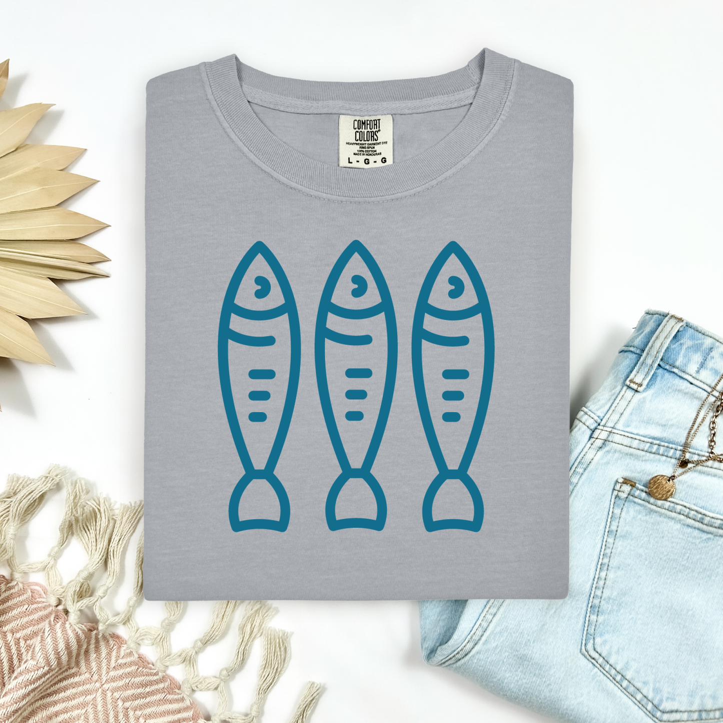 Comfort Colors 1717 Fisherman's Aesthetic t shirt with three sardines in a  one color graphic. Picture on a flay lay mockup with pair of blue jeans and blanket and a wicker sun shaped accessory.