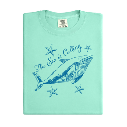 Comfort Colors t-shirt featuring a blue whale illustration surrounded by starfish and the handwritten-style text “The Sea is Calling.” The detailed marine artwork creates a calming coastal aesthetic. This unisex garment-dyed tee offers a soft vintage feel and relaxed fit, perfect for ocean lovers, beach trips, and anyone who enjoys sea-themed graphic shirts with a coastal vibe.