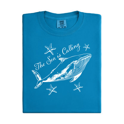 Comfort Colors t-shirt featuring a blue whale illustration surrounded by starfish and the handwritten-style text “The Sea is Calling.” The detailed marine artwork creates a calming coastal aesthetic. This unisex garment-dyed tee offers a soft vintage feel and relaxed fit, perfect for ocean lovers, beach trips, and anyone who enjoys sea-themed graphic shirts with a coastal vibe.