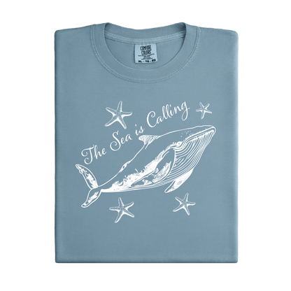 Comfort Colors t-shirt featuring a blue whale illustration surrounded by starfish and the handwritten-style text “The Sea is Calling.” The detailed marine artwork creates a calming coastal aesthetic. This unisex garment-dyed tee offers a soft vintage feel and relaxed fit, perfect for ocean lovers, beach trips, and anyone who enjoys sea-themed graphic shirts with a coastal vibe.