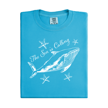 Comfort Colors t-shirt featuring a blue whale illustration surrounded by starfish and the handwritten-style text “The Sea is Calling.” The detailed marine artwork creates a calming coastal aesthetic. This unisex garment-dyed tee offers a soft vintage feel and relaxed fit, perfect for ocean lovers, beach trips, and anyone who enjoys sea-themed graphic shirts with a coastal vibe.