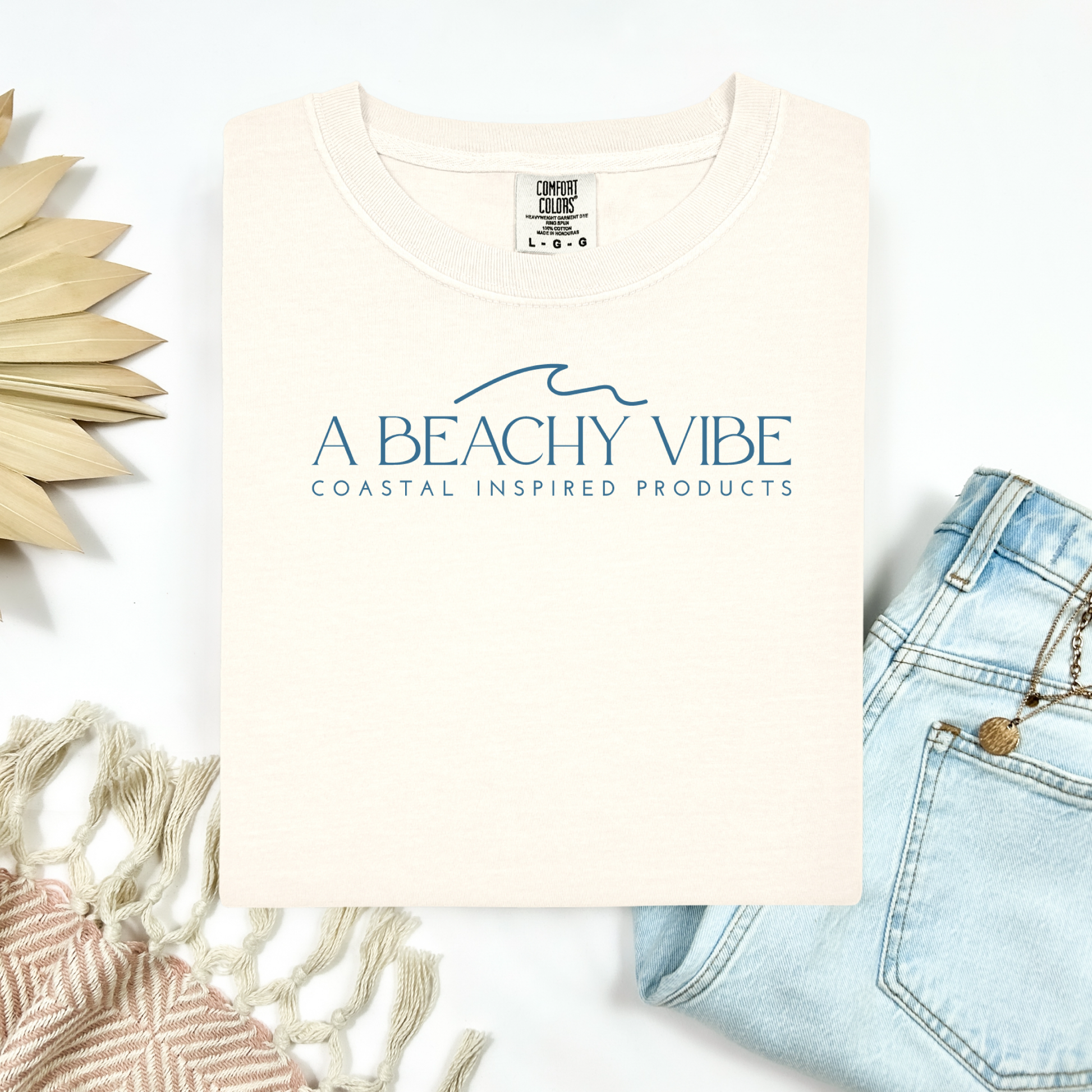 Comfort Colors 1717 "A Beachy Vibe" t shirt with a simple wave one color graphic. Picture on a flay lay mockup with pair of blue jeans and blanket and a wicker sun shaped accessory.
