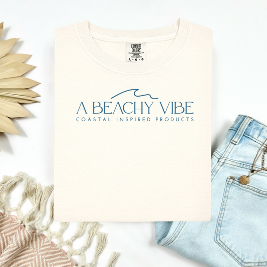 Comfort Colors 1717 "A Beachy Vibe" t shirt with a simple wave one color graphic. Picture on a flay lay mockup with pair of blue jeans and blanket and a wicker sun shaped accessory.