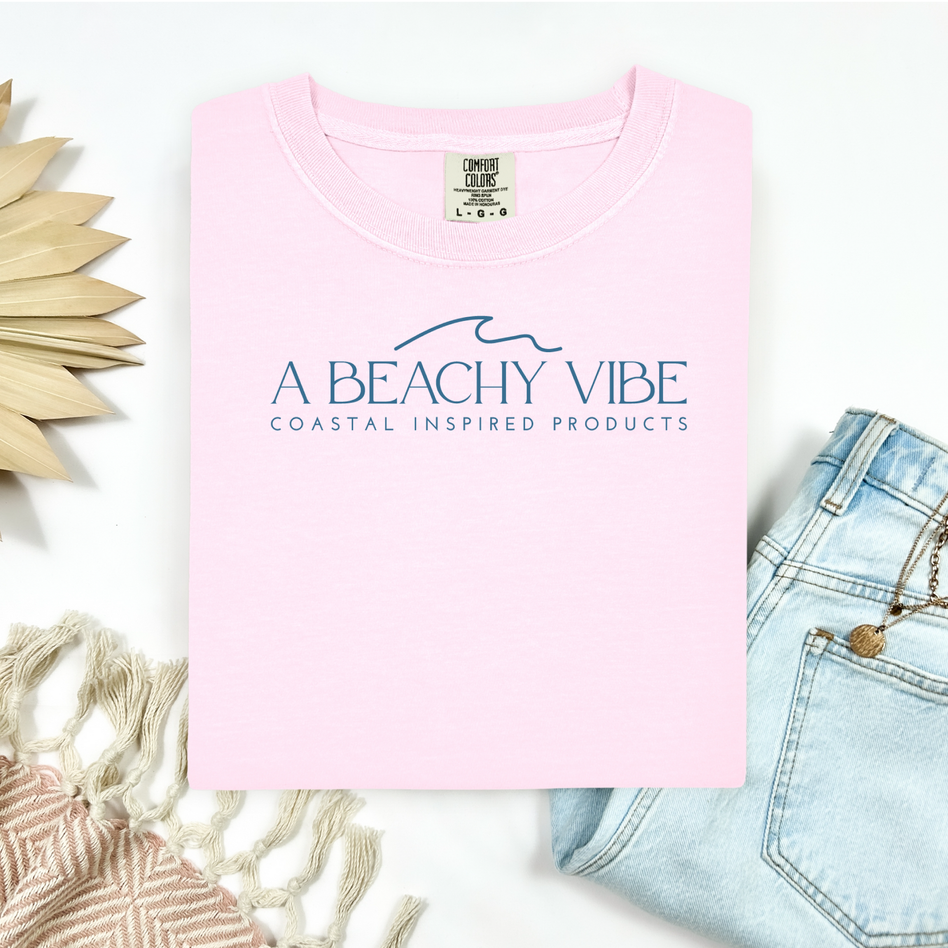 Comfort Colors 1717 "A Beachy Vibe" t shirt with a simple wave one color graphic. Picture on a flay lay mockup with pair of blue jeans and blanket and a wicker sun shaped accessory.