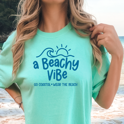 Person wearing a light blue t-shirt with 'a Beachy Vibe' text and graphics on a beach.