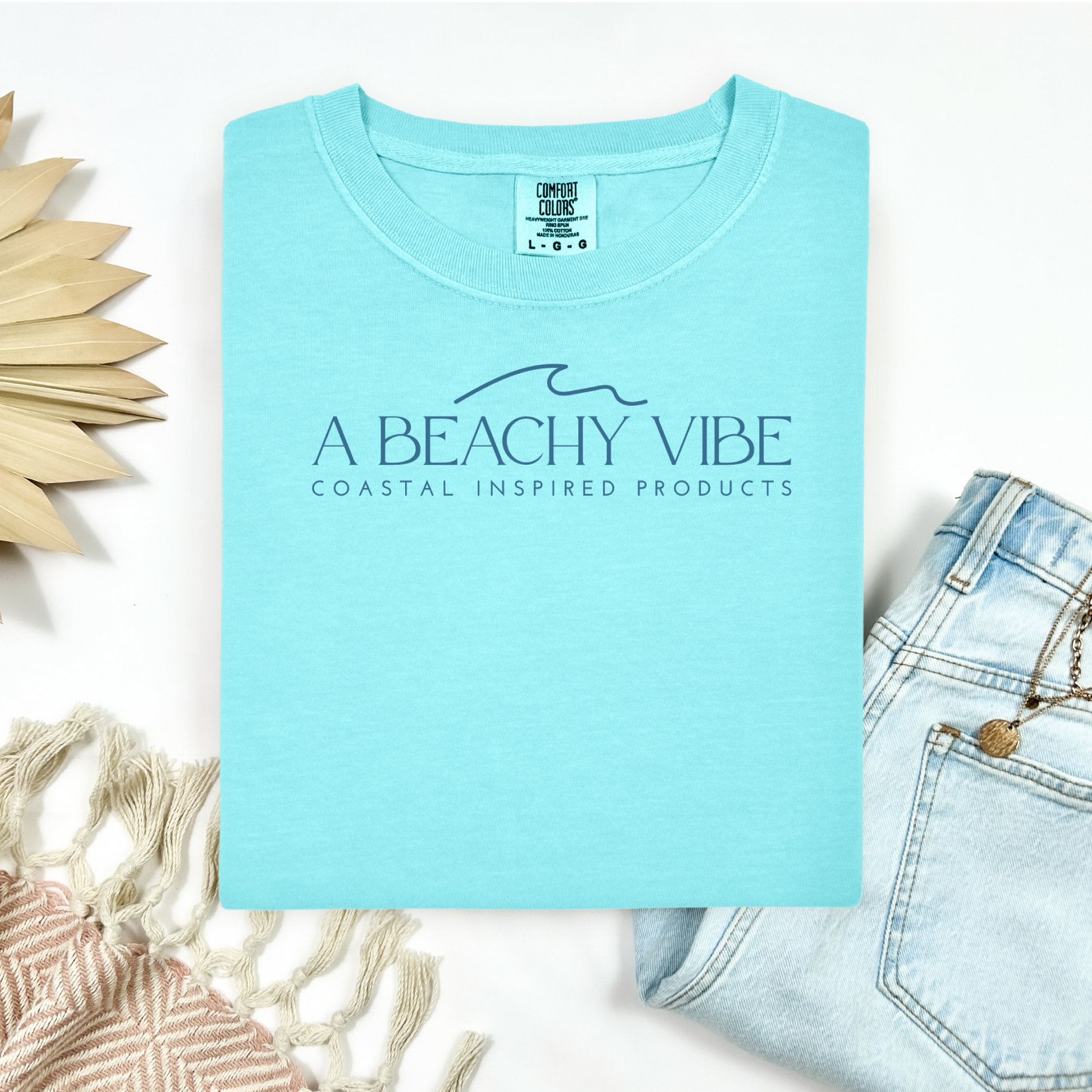 Comfort Colors 1717 "A Beachy Vibe" t shirt with a simple wave one color graphic. Picture on a flay lay mockup with pair of blue jeans and blanket and a wicker sun shaped accessory.