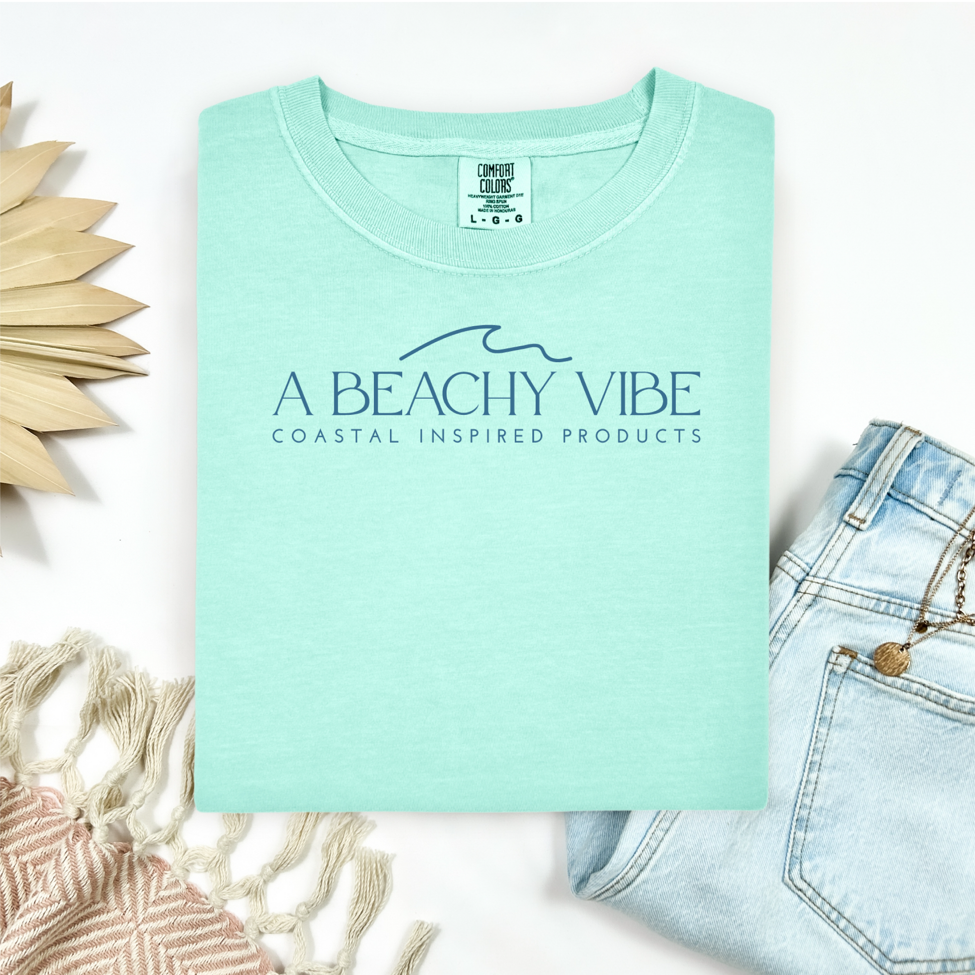 Comfort Colors 1717 "A Beachy Vibe" t shirt with a simple wave one color graphic. Picture on a flay lay mockup with pair of blue jeans and blanket and a wicker sun shaped accessory.