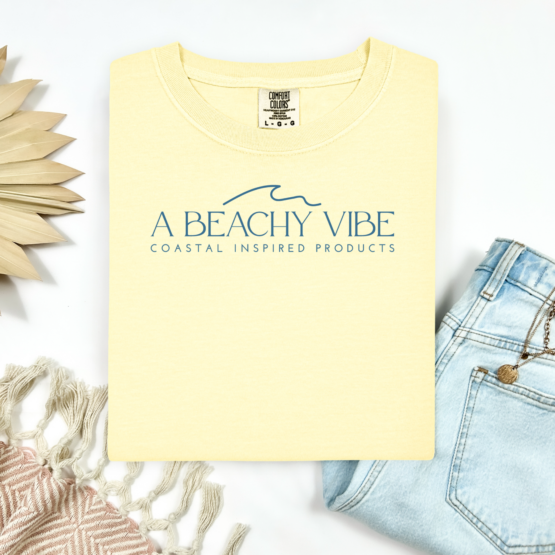 Comfort Colors 1717 "A Beachy Vibe" t shirt with a simple wave one color graphic. Picture on a flay lay mockup with pair of blue jeans and blanket and a wicker sun shaped accessory.