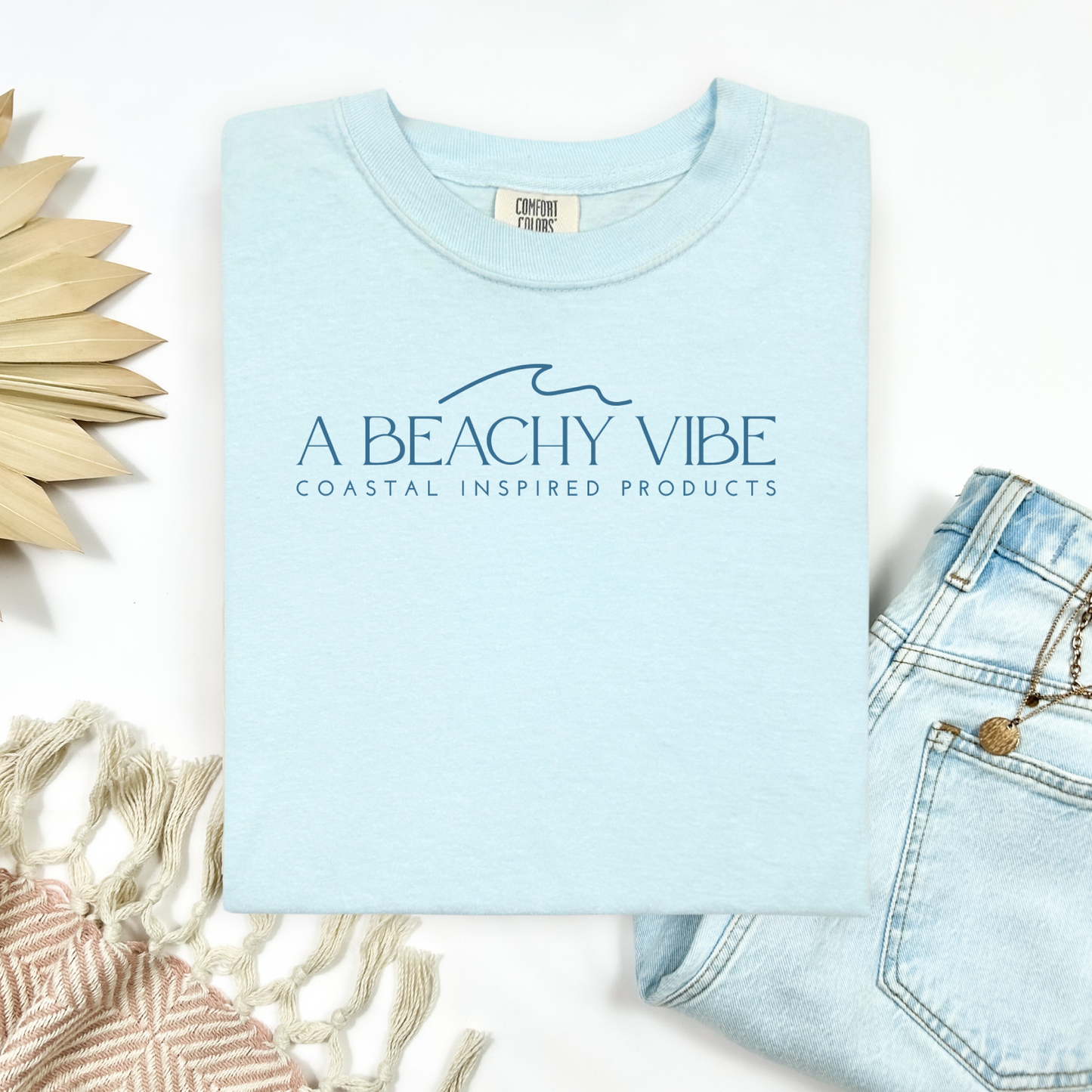 Comfort Colors 1717 "A Beachy Vibe" t shirt with a simple wave one color graphic. Picture on a flay lay mockup with pair of blue jeans and blanket and a wicker sun shaped accessory.