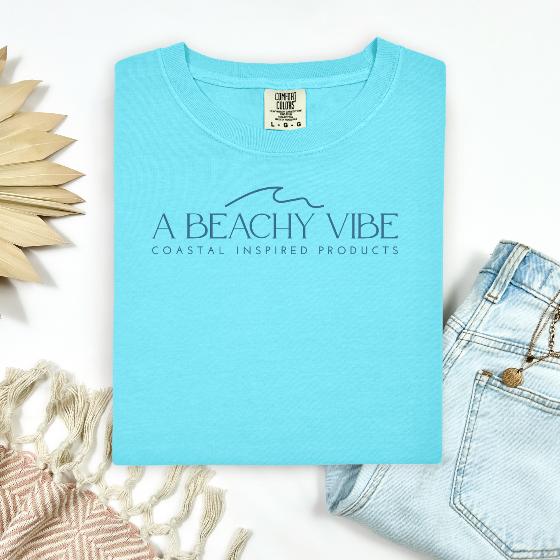 Comfort Colors 1717 "A Beachy Vibe" t shirt with a simple wave one color graphic. Picture on a flay lay mockup with pair of blue jeans and blanket and a wicker sun shaped accessory.