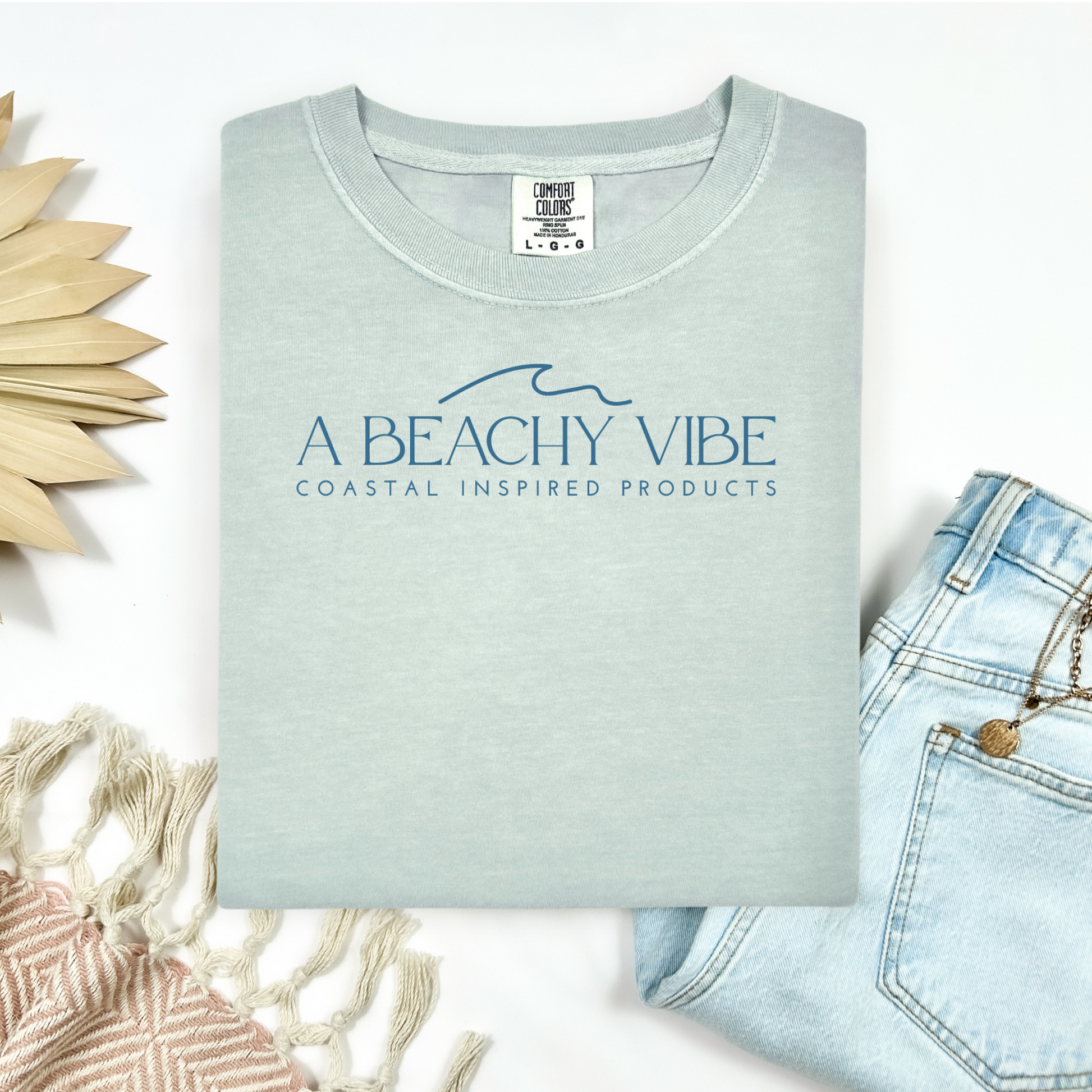 Comfort Colors 1717 "A Beachy Vibe" t shirt with a simple wave one color graphic. Picture on a flay lay mockup with pair of blue jeans and blanket and a wicker sun shaped accessory.