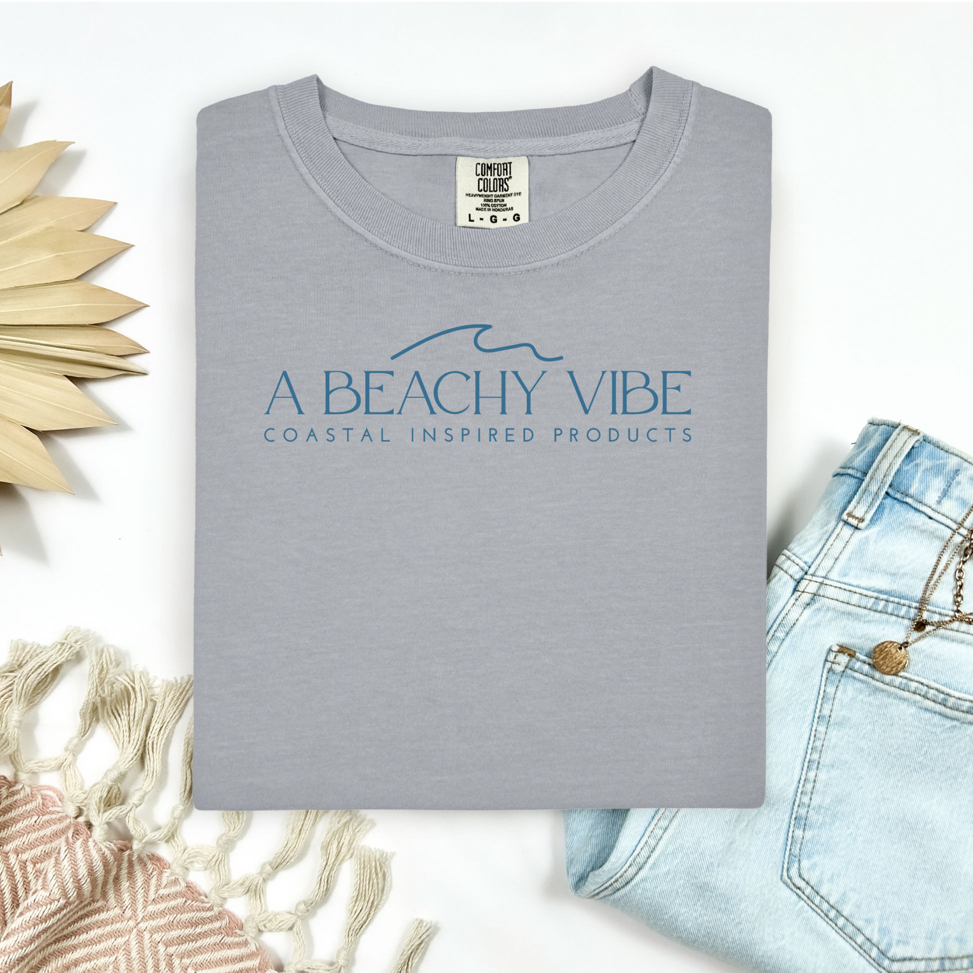 Comfort Colors 1717 "A Beachy Vibe" t shirt with a simple wave one color graphic. Picture on a flay lay mockup with pair of blue jeans and blanket and a wicker sun shaped accessory.