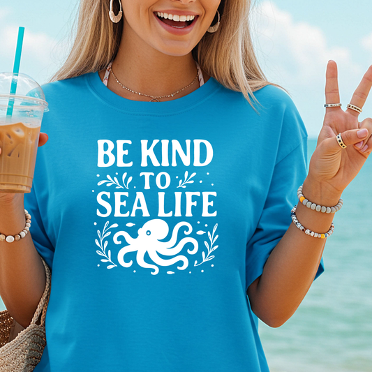 Person wearing a blue t-shirt with 'Be Kind to Sea Life' and an octopus graphic, holding a drink by the beach.