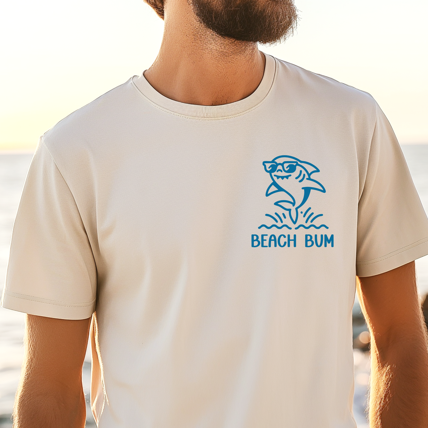 Person wearing a beige t-shirt with a blue shark graphic and 'Beach Bum' text at the beach.