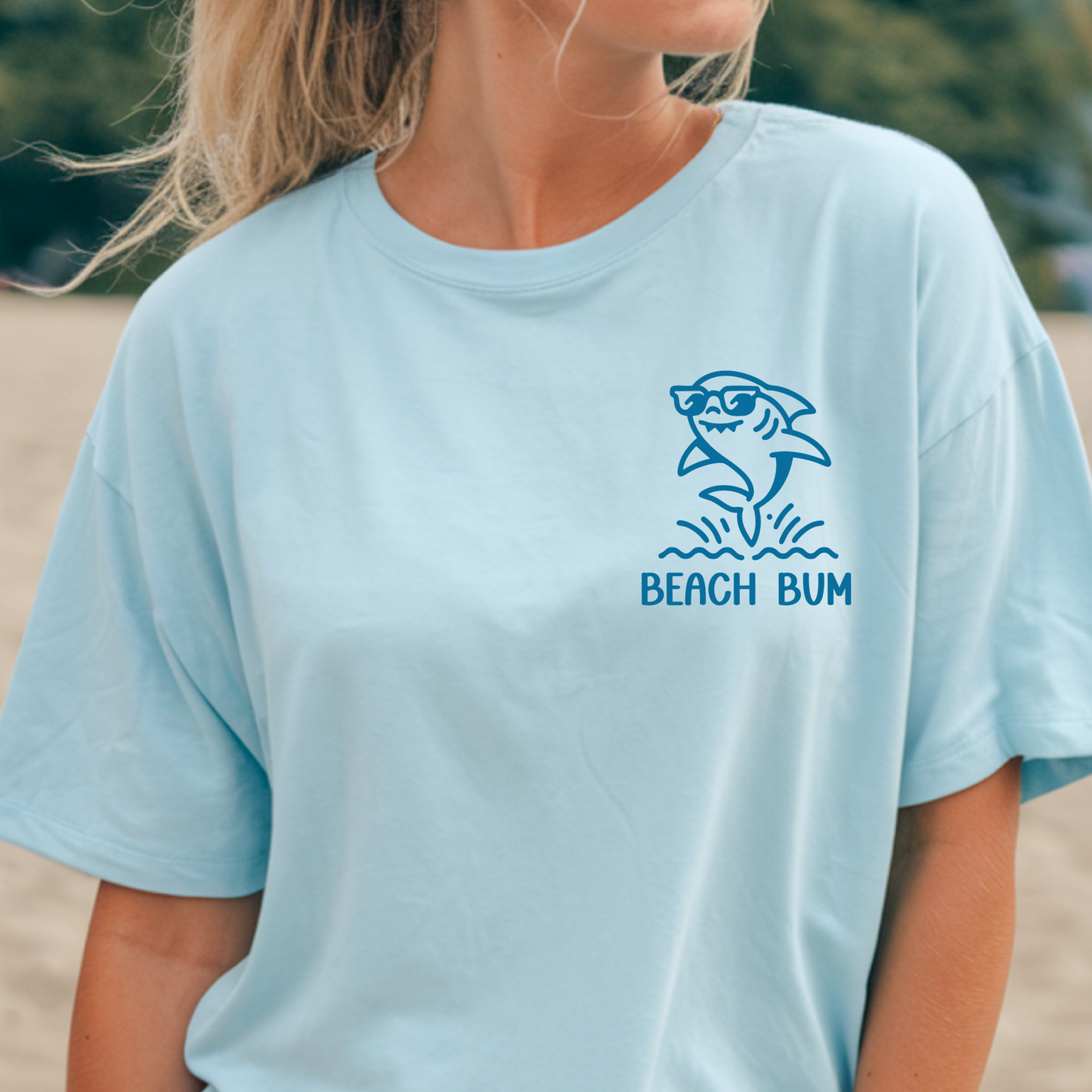 Person wearing a light blue t-shirt with a shark graphic and 'Beach Bum' text on a beach.