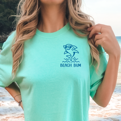 Person wearing a green t-shirt with a shark graphic and 'Beach Bum' text at a beach.