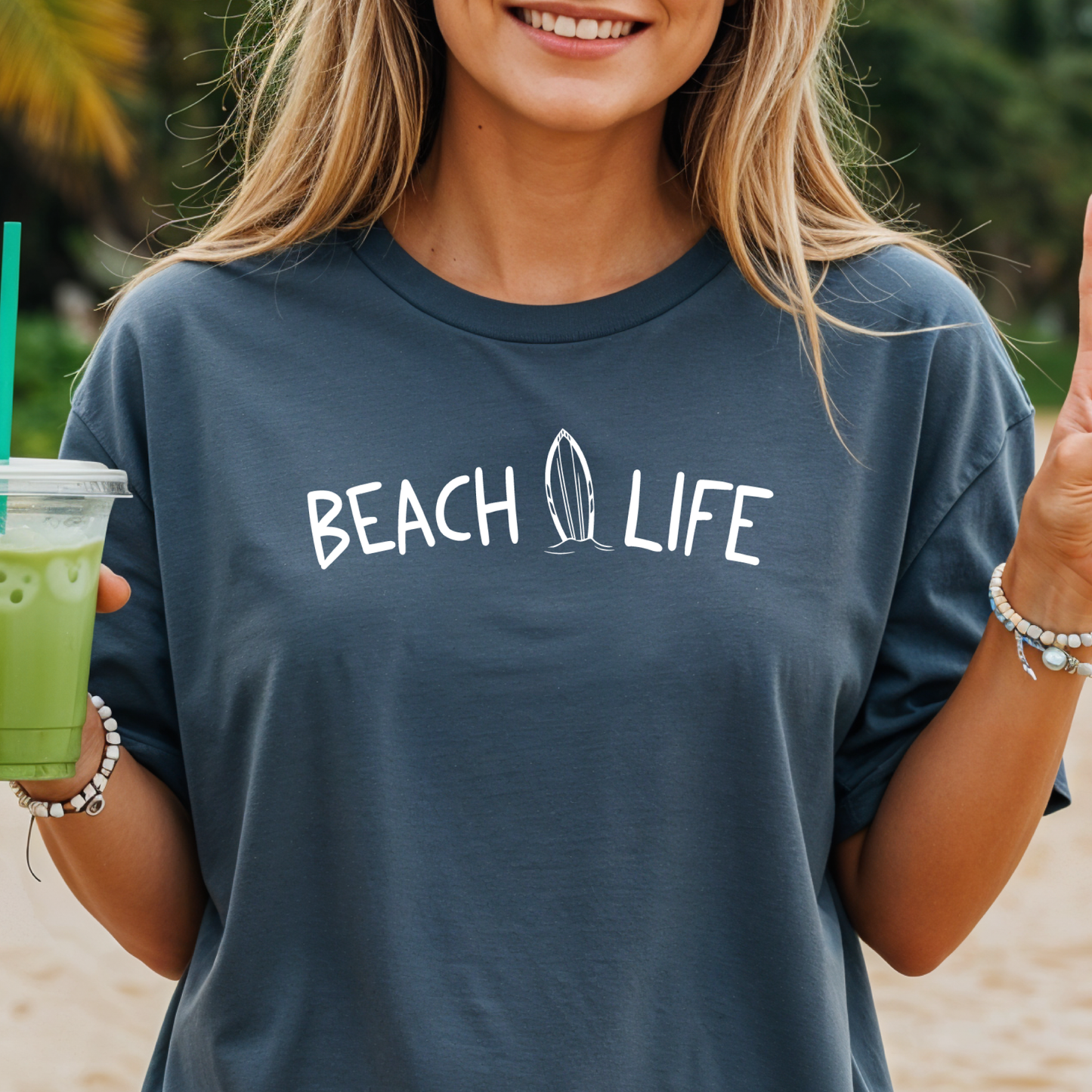 Woman wearing a 'Beach Life' t-shirt holding a green drink on a beach.