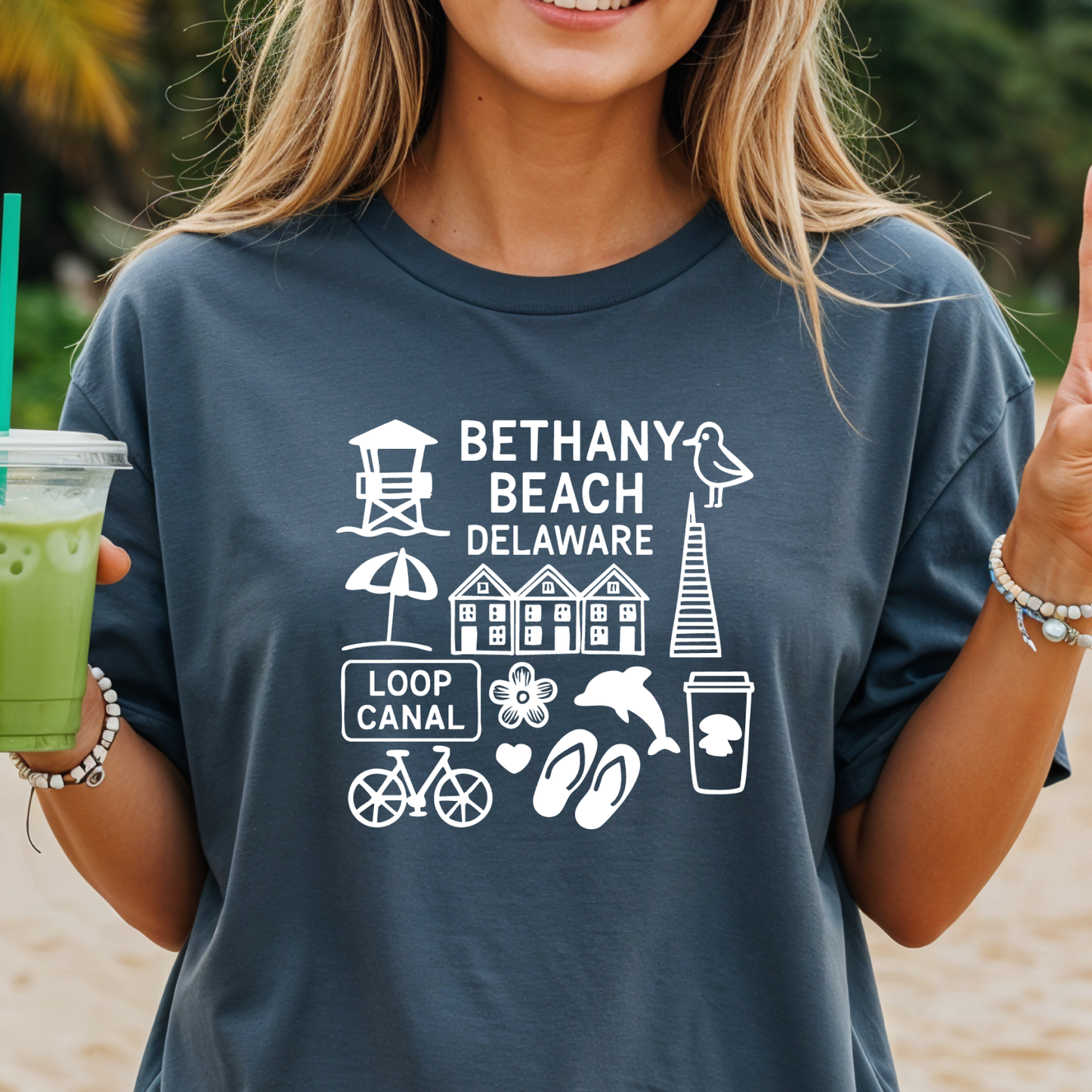 Person wearing a navy blue t-shirt with Bethany Beach, Delaware graphics, holding a green drink.