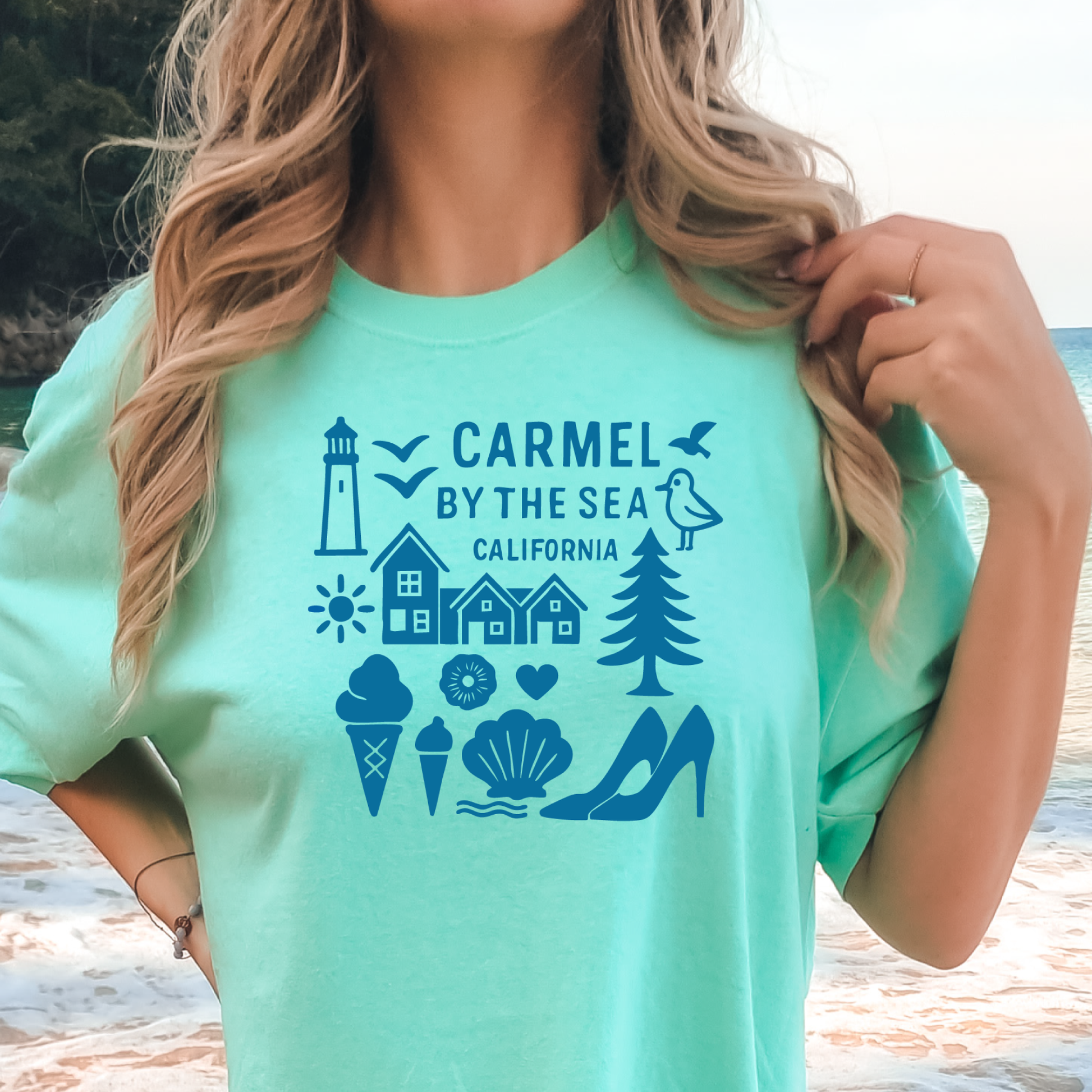 Person wearing a teal t-shirt with 'Carmel by the Sea, California' design on a beach.