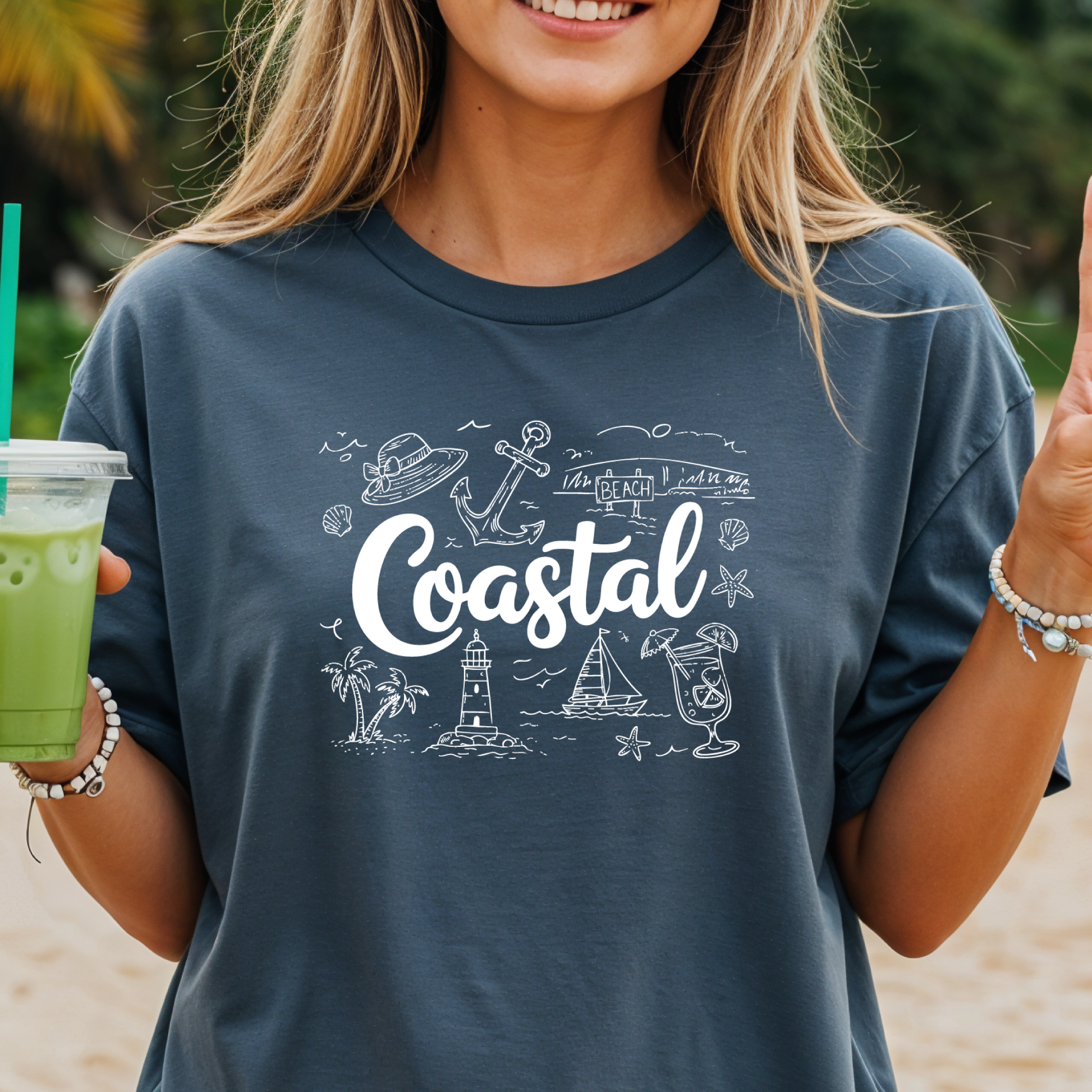 Person wearing a blue denim t-shirt with 'Coastal' design and holding a drink on a beach.