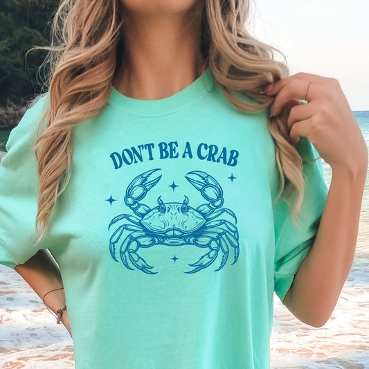 Person wearing a light green t-shirt with a crab graphic and text on a beach.