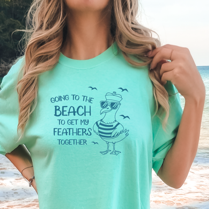 Comfort Colors t-shirt featuring a humorous blue seagull graphic wearing sunglasses and a striped shirt, beside the text “Going to the Beach to Get My Feathers Together.” Several flying birds surround the design, adding a fun coastal feel. This unisex garment-dyed tee offers a soft, vintage texture and relaxed fit, perfect for beach lovers, ocean fans, and anyone who enjoys playful summer graphic shirts.