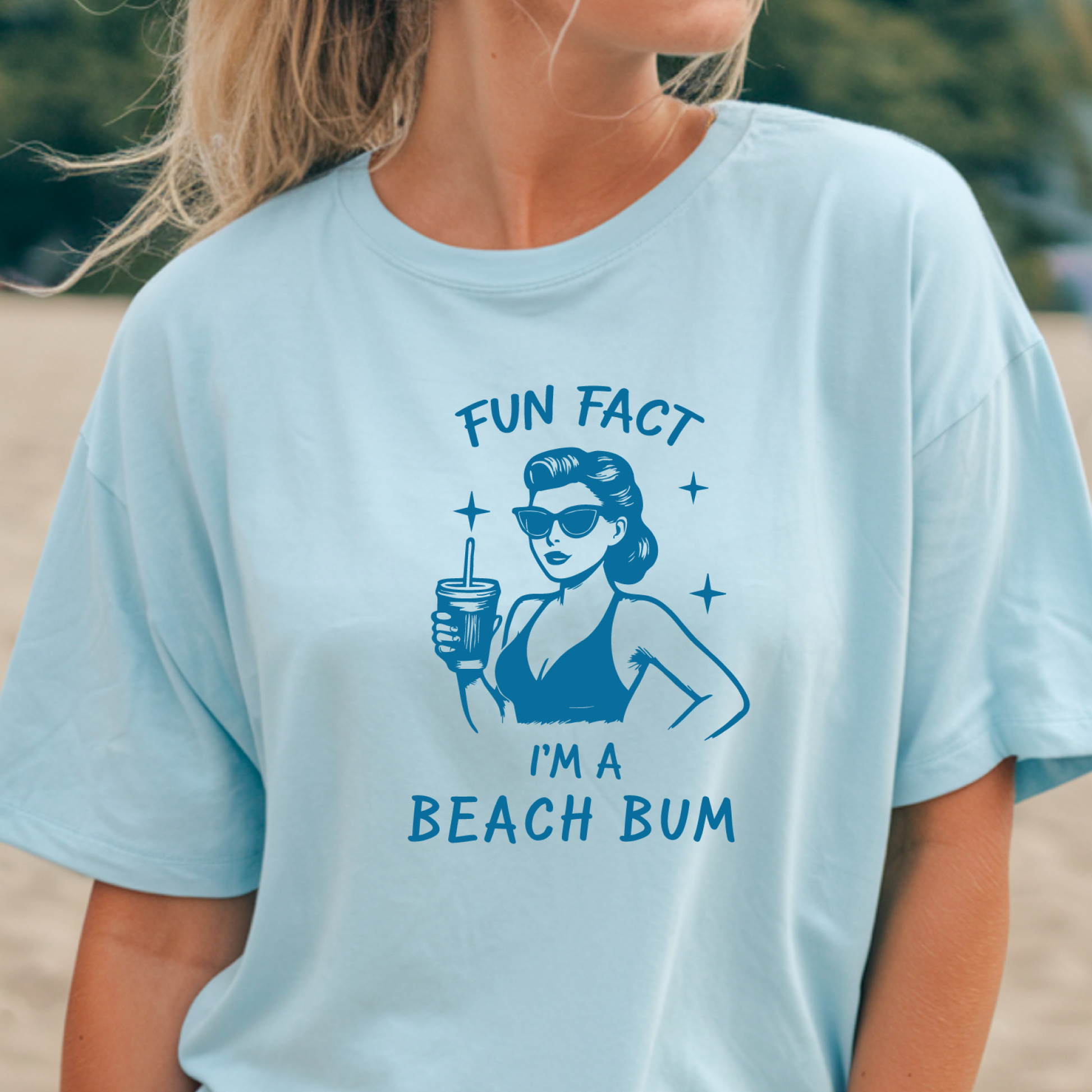 Light blue t-shirt with a graphic and text 'Fun Fact I'm a Beach Bum' worn by a person on a beach.