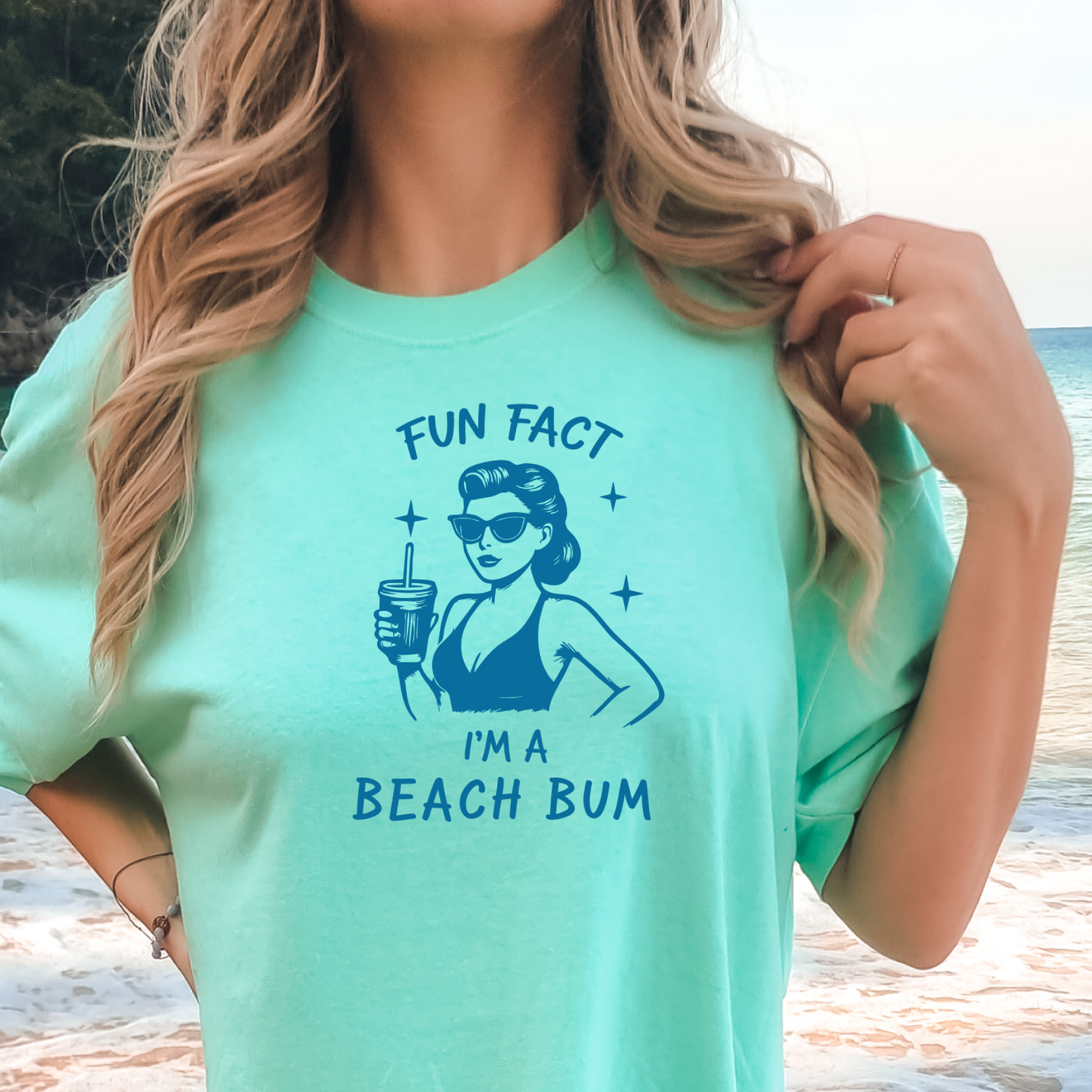 Person wearing a light green t-shirt with a graphic and text 'Fun Fact I'm a Beach Bum' at the beach.