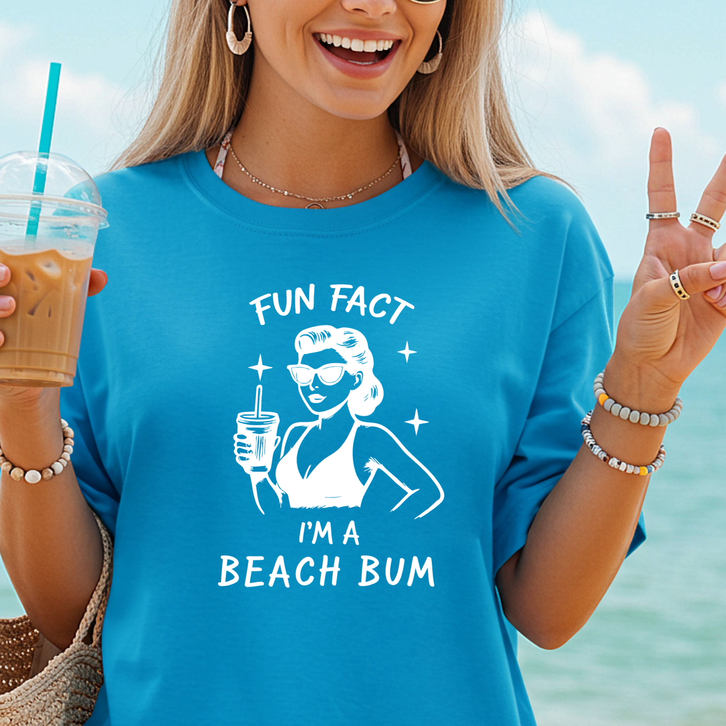Woman wearing a blue t-shirt with 'Fun Fact: I'm a Beach Bum' design, holding a drink and making a peace sign.
