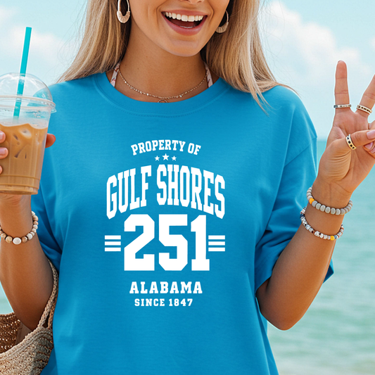 Person wearing a blue t-shirt with 'Property of Gulf Shores 251 Alabama Since 1847' text, holding a drink by the beach.