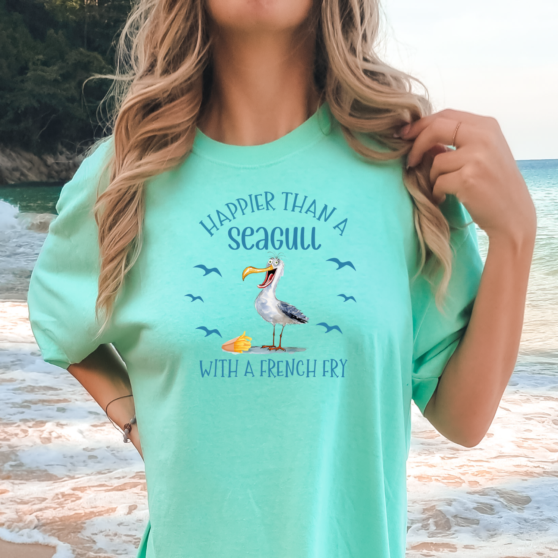 Woman wearing a mint green t-shirt with a seagull graphic and text on a beach.