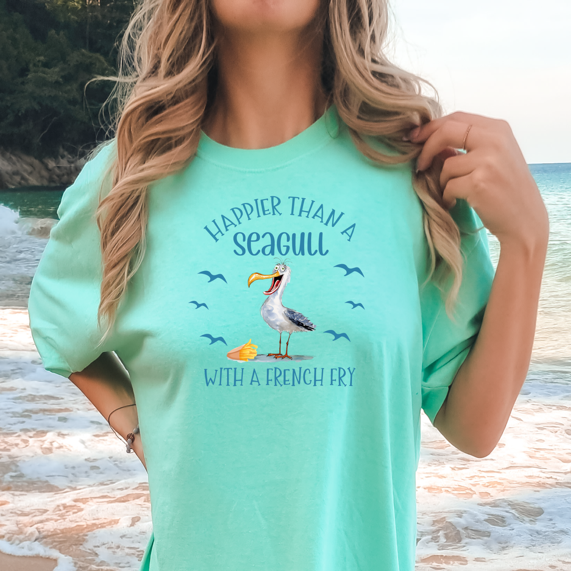 Woman wearing a mint green t-shirt with a seagull graphic and text on a beach.
