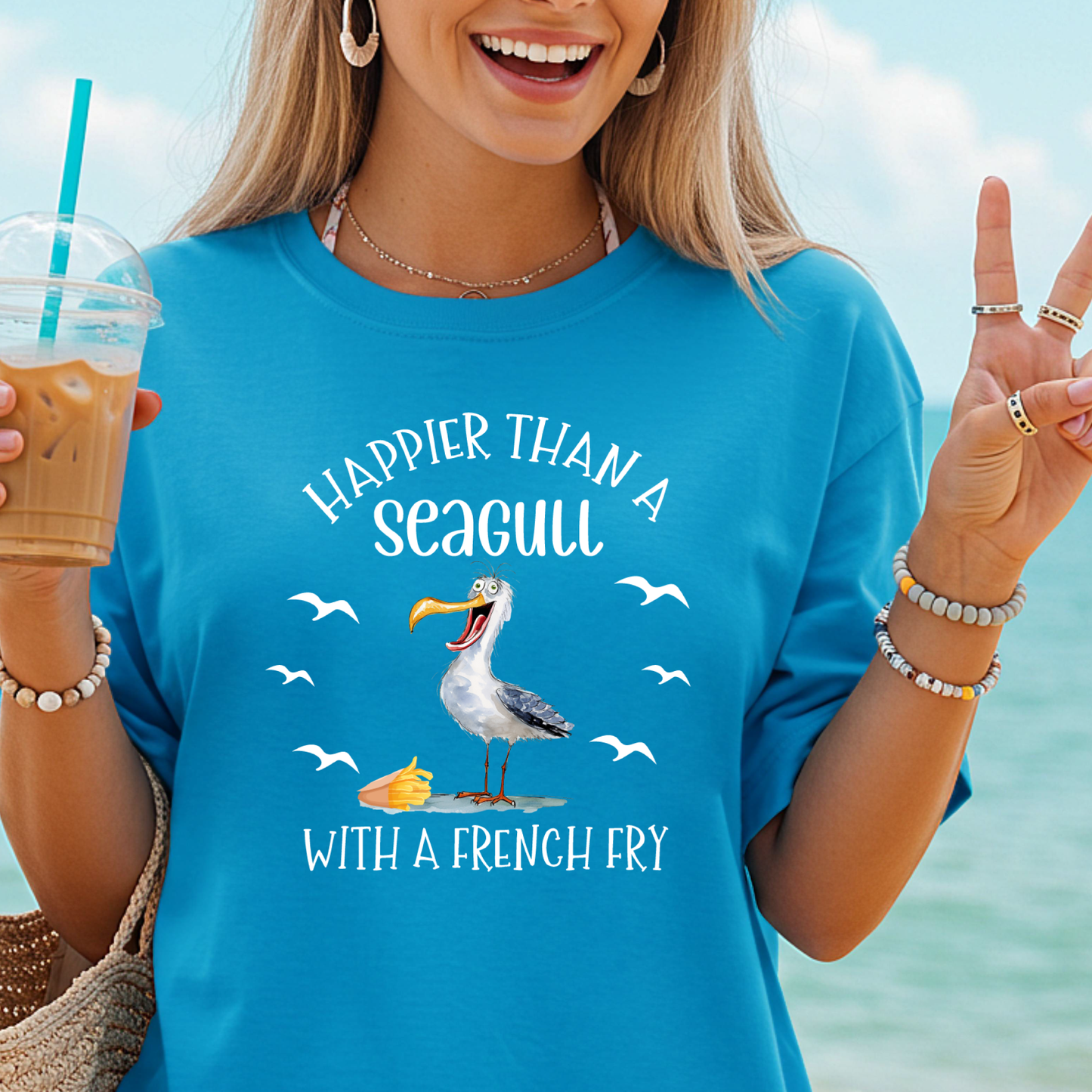 Woman wearing a blue t-shirt with a seagull graphic and text, holding a drink by the beach.