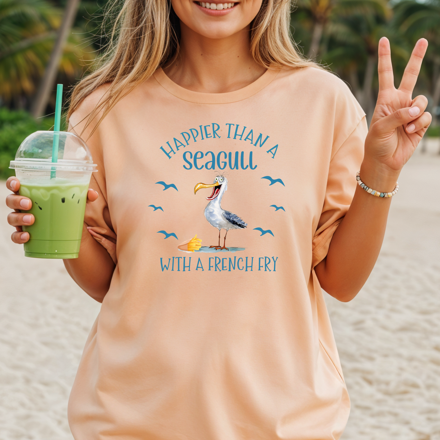 Woman on a beach wearing a peach t-shirt with a seagull graphic and text, holding a drink.