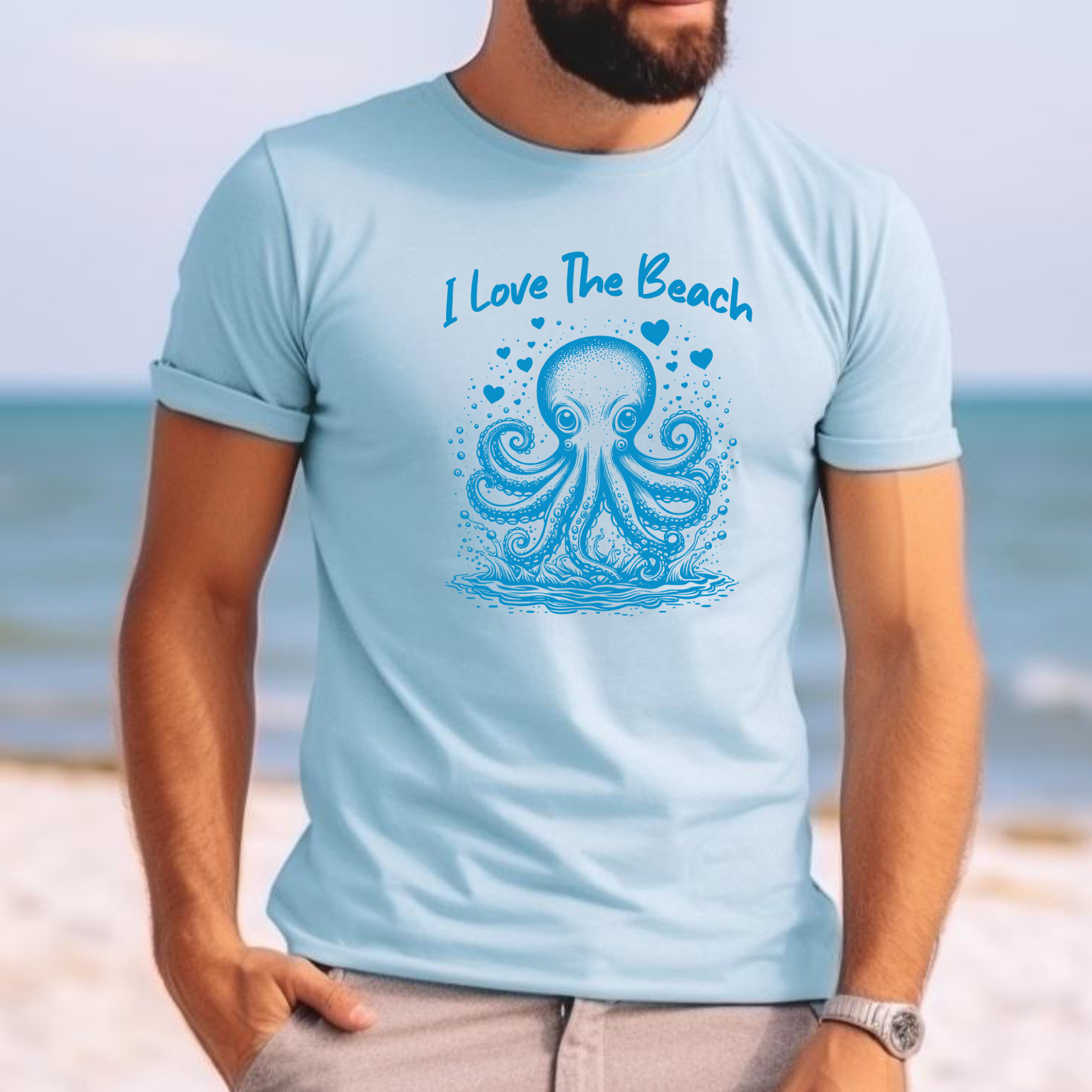 The Comfort Colors 1717 "I Love The Beach" T-shirt features a playful one-color graphic of an octopus surrounded by hearts, capturing a fun and beach-loving spirit. Worn relaxed fit by a man standing on the beach, the tee embodies relaxed coastal vibes and effortless summer style, perfect for sunny days by the shore.