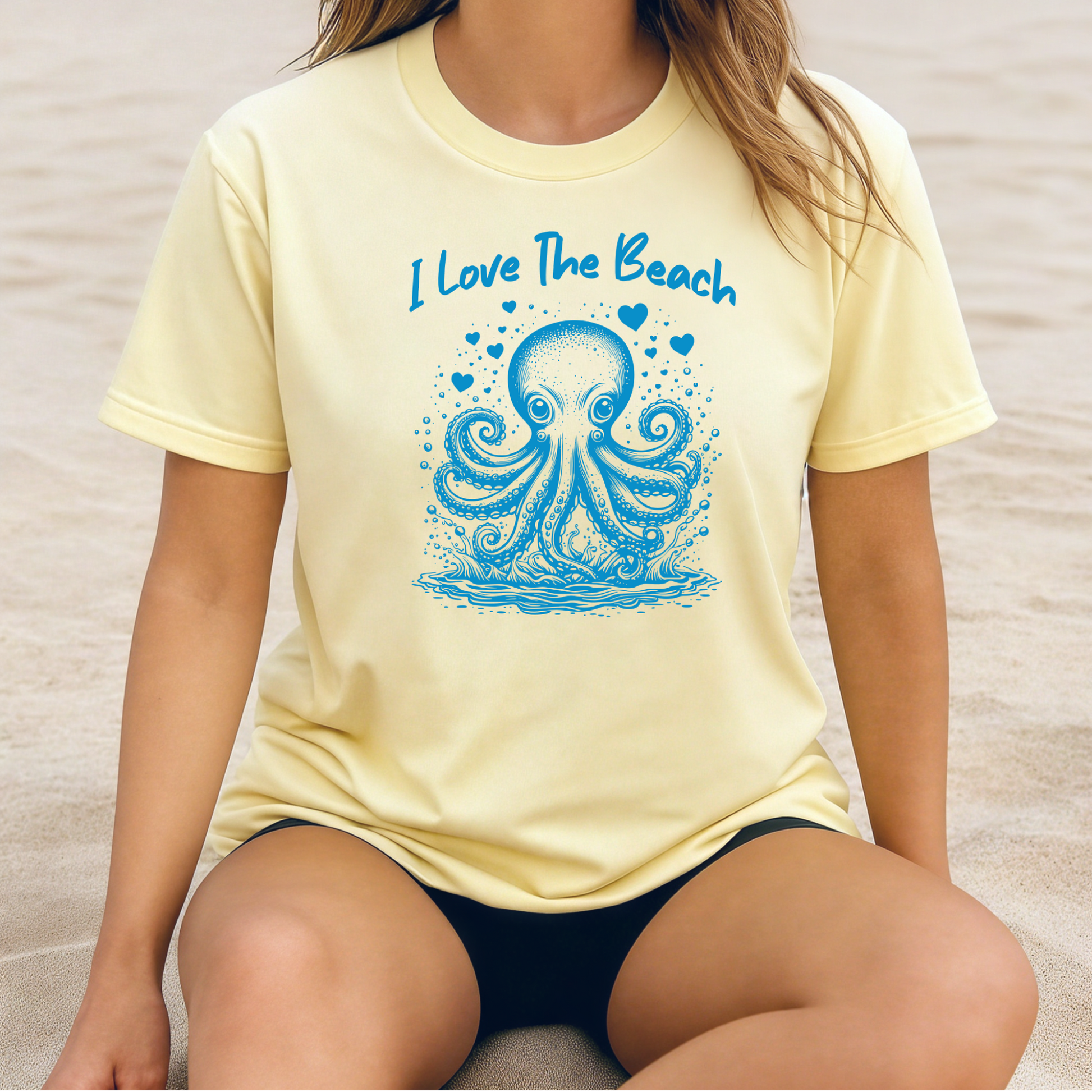 The Comfort Colors 1717 "I Love The Beach" T-shirt features a playful one-color graphic of an octopus surrounded by hearts, capturing a fun and beach-loving spirit. Worn oversized by a girl sitting on the beach, the tee embodies relaxed coastal vibes and effortless summer style, perfect for sunny days by the shore.