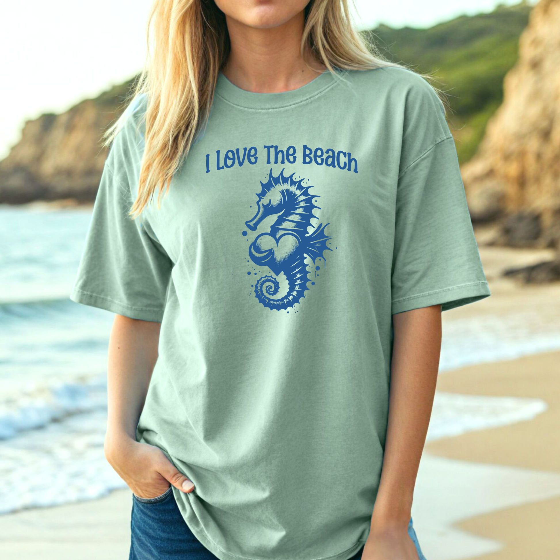 Comfort Colors 1717 "I Love The Beach" t shirt with a seahorse holding a heart graphics in one color. Picture of a girl in an oversize shirt standing on the beach. 