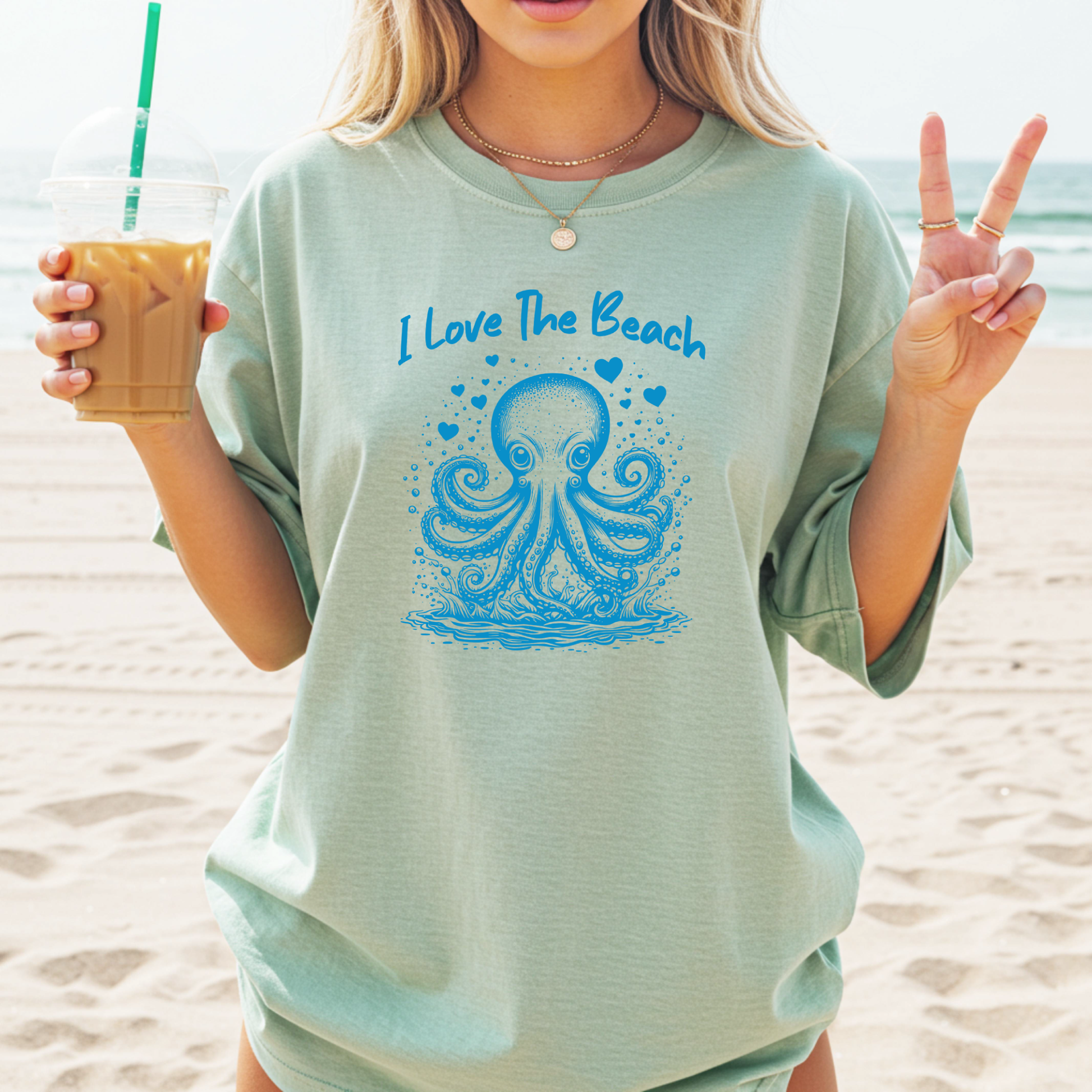 The Comfort Colors 1717 "I Love The Beach" T-shirt features a playful one-color graphic of an octopus surrounded by hearts, capturing a fun and beach-loving spirit. Worn oversized by a girl standing on the beach, the tee embodies relaxed coastal vibes and effortless summer style, perfect for sunny days by the shore.