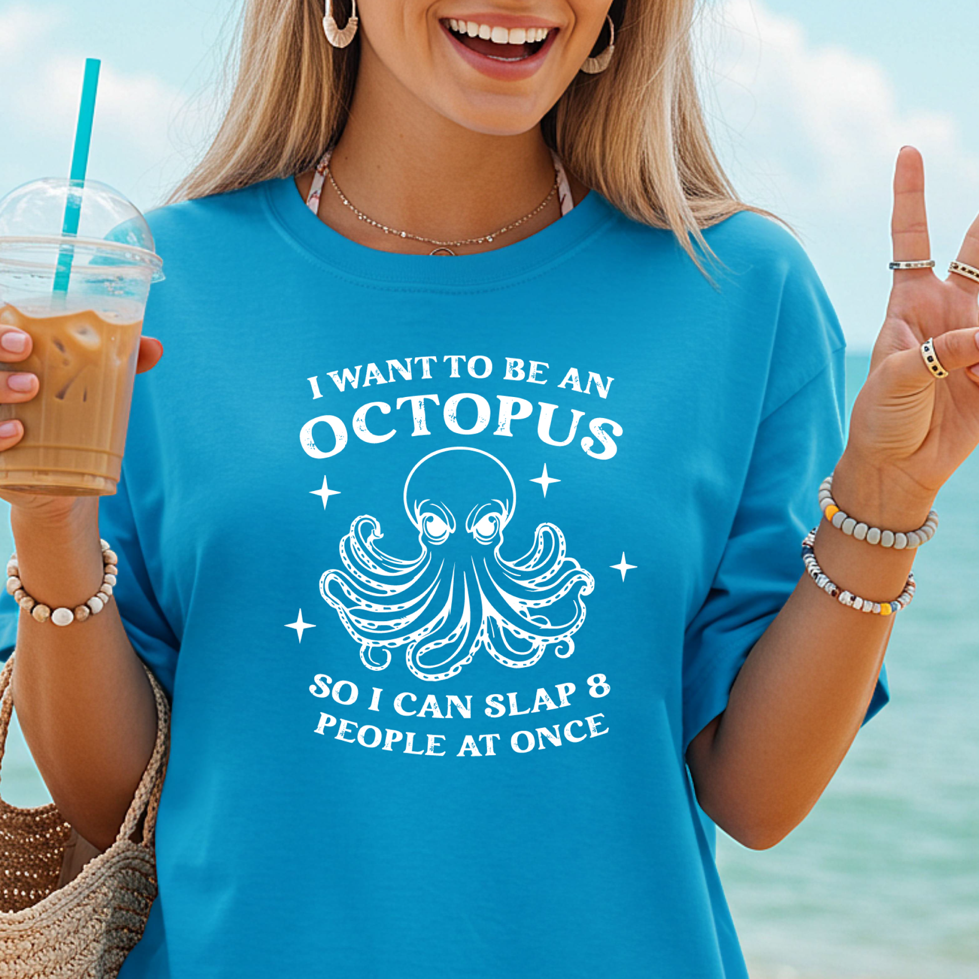 Person wearing a blue sapphire t-shirt with an octopus graphic and text, holding a drink by the beach.