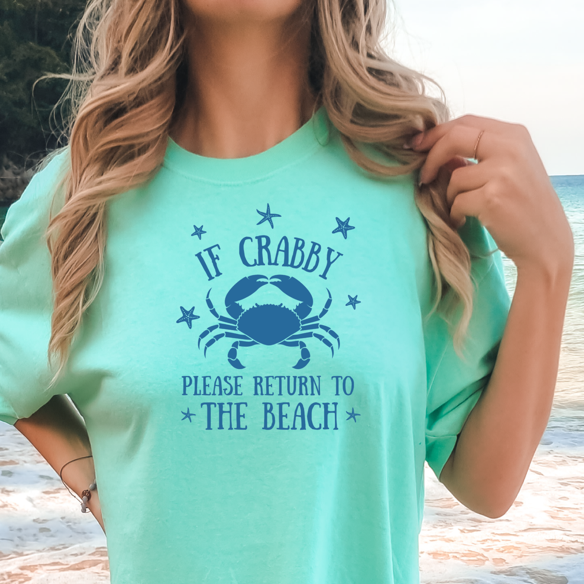 Person wearing a light green t-shirt with a crab graphic and text at the beach.