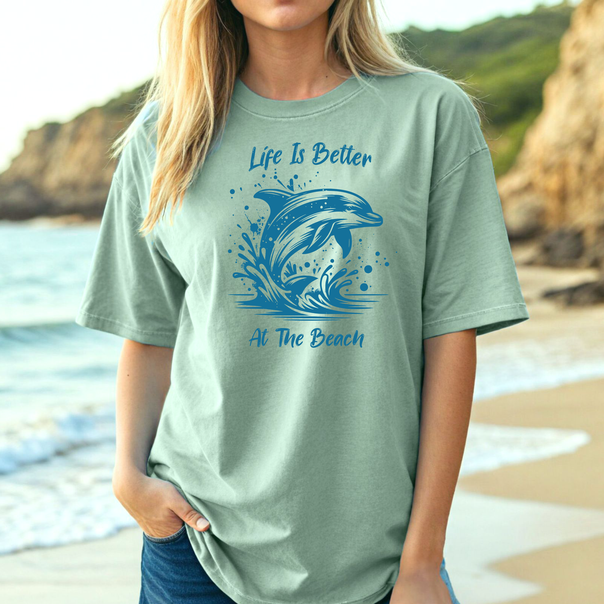 Comfort Colors 1717 "Life is Better at the Beach" t shirt with a dolphin jumping out of the water in one color graphic. Picture of a girl on the beach in an oversized t shirt.