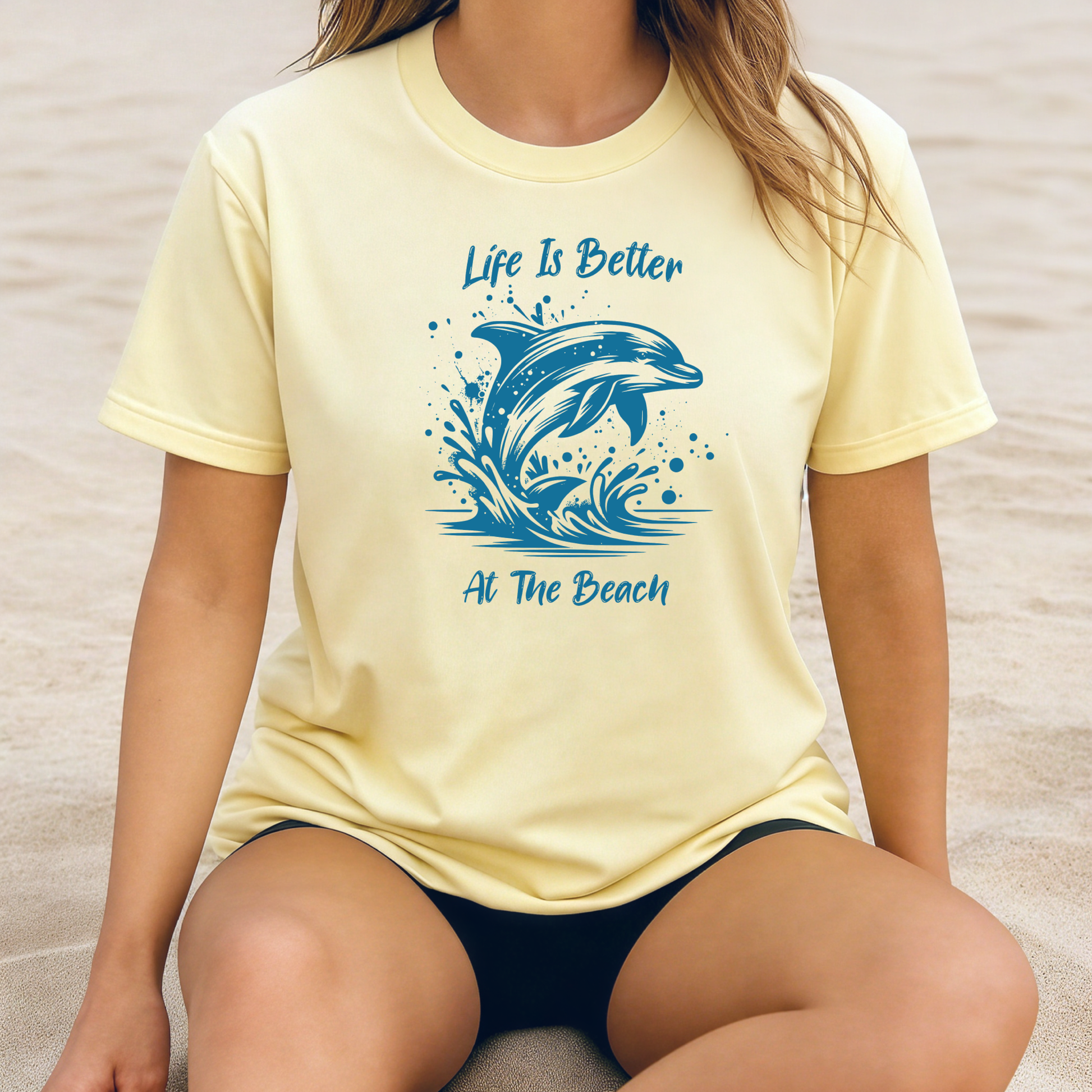 Comfort Colors 1717 "Life is Better at the Beach" t shirt with a dolphin jumping out of the water in one color graphic. Picture of a girl sitting on the beach in an oversized t shirt.