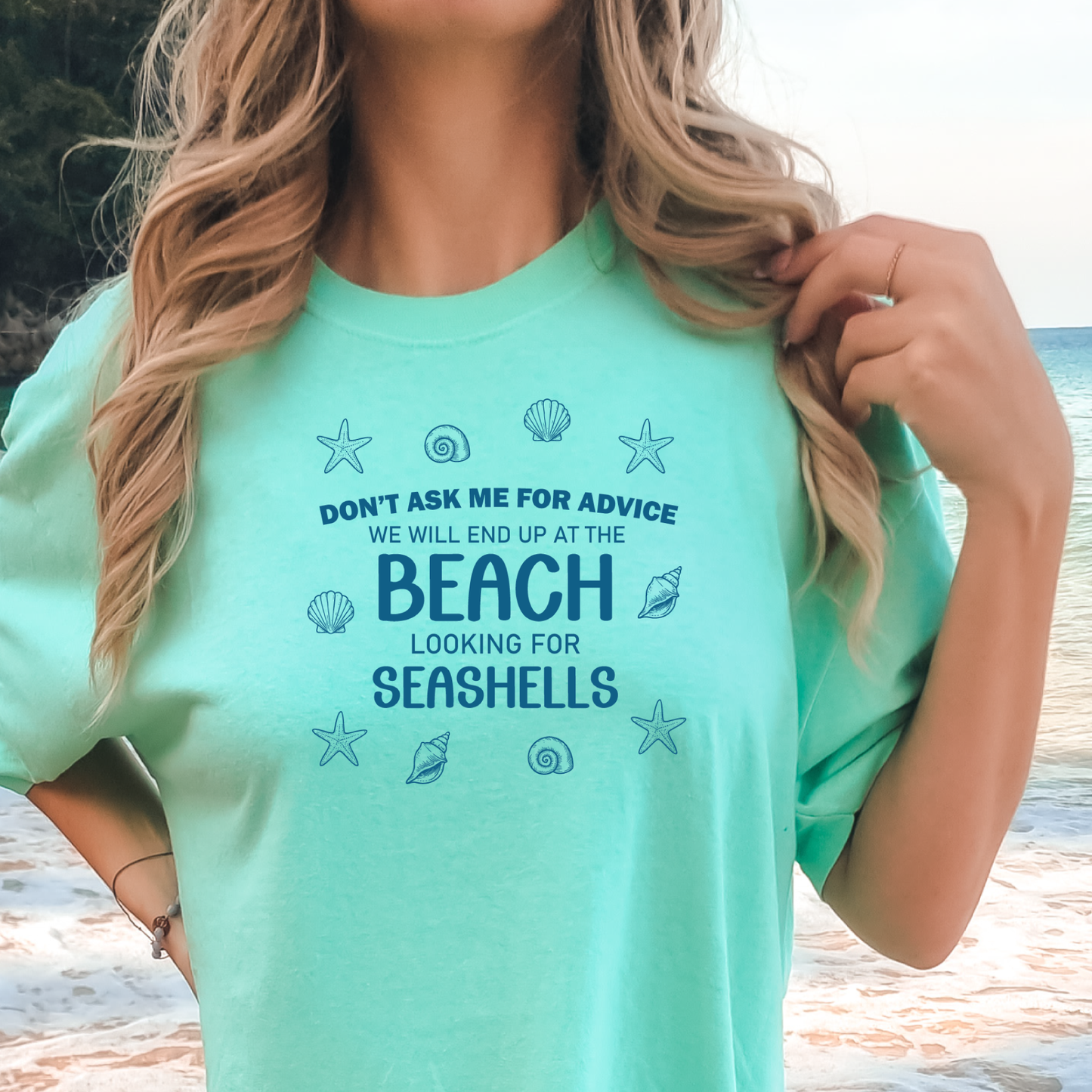 Person wearing a light green t-shirt with beach-themed text and graphics, standing by a beach.