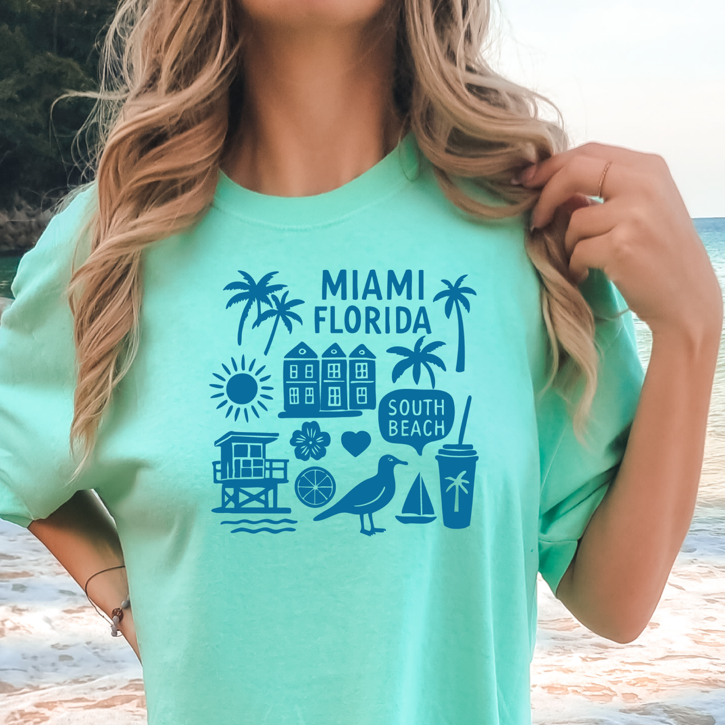 Person wearing a green t-shirt with 'Miami Florida' and beach icons on a beach background